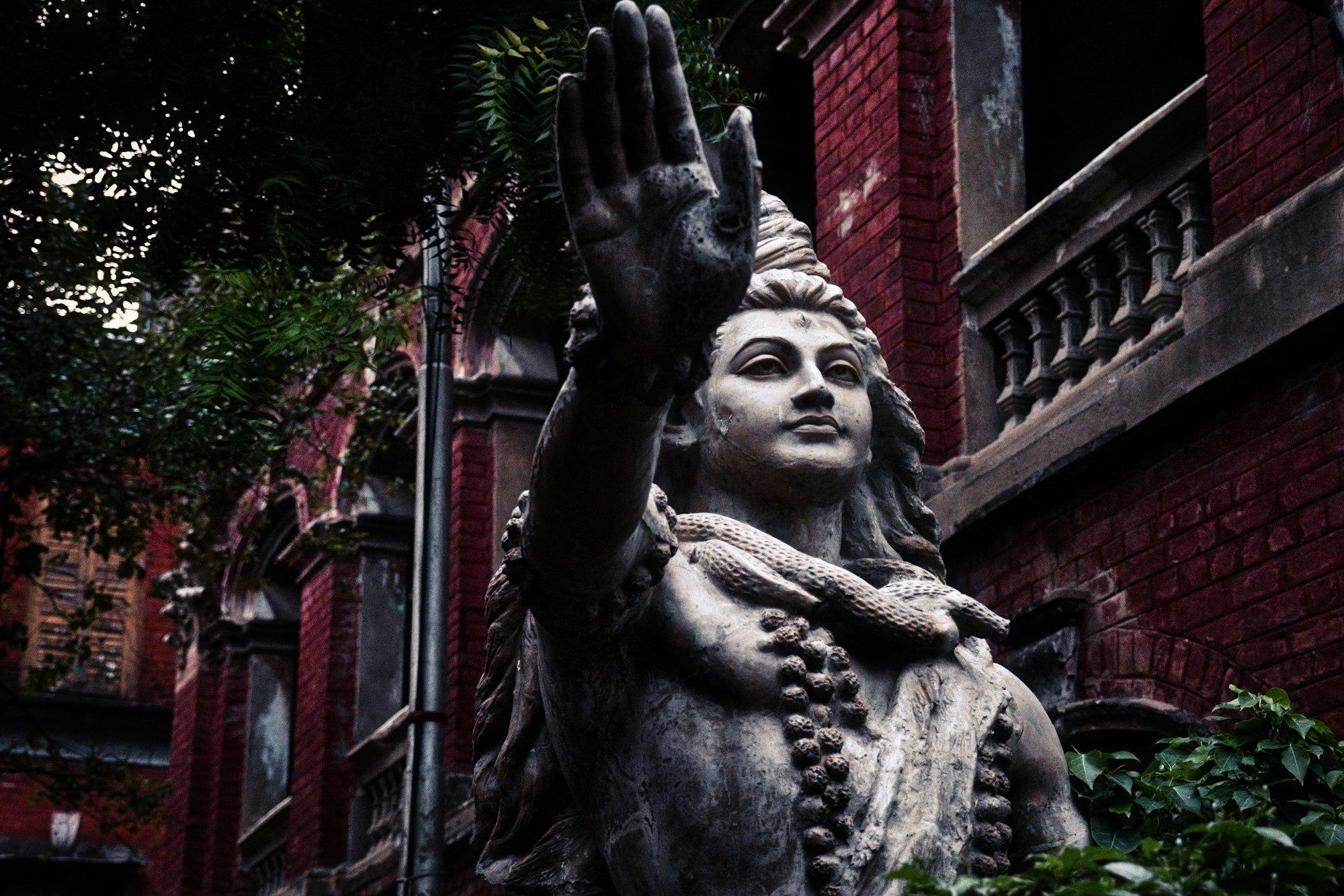 A weathered stone deity statue in a Kolkata alley beside a red colonial-era building