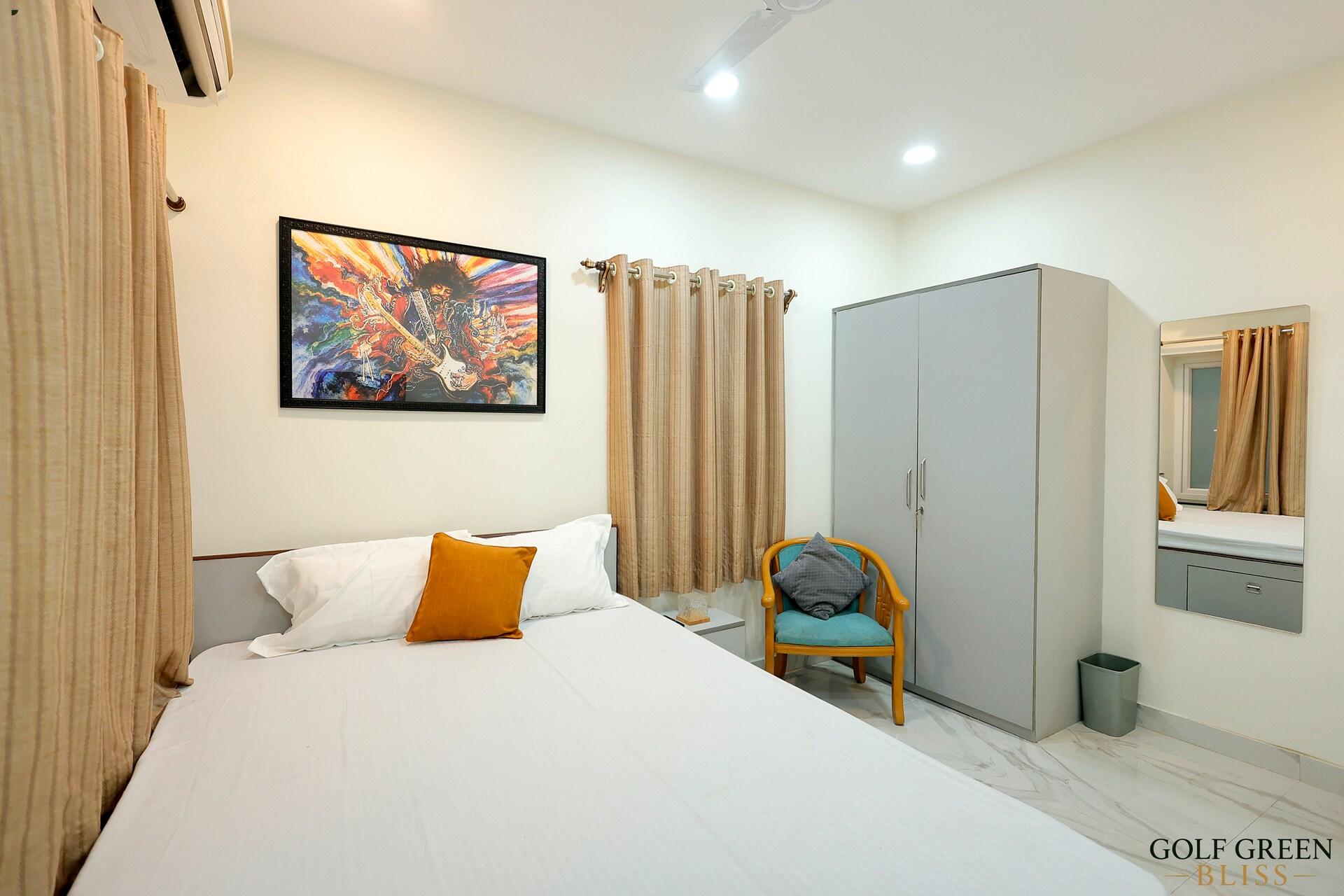 Bedroom one with a queen bed, framed artwork, a wardrobe and an armchair beside the curtained window